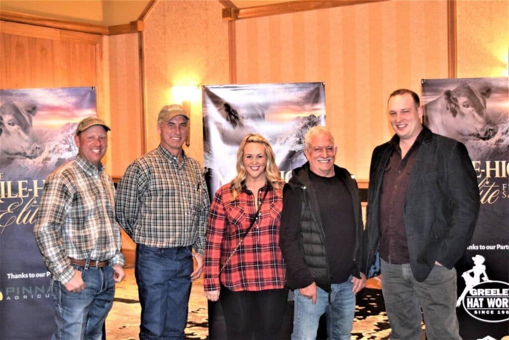 Five people stand smiling indoors in front of two banners featuring horses and event logos. Three men wear casual jackets and plaid shirts, while a woman in the middle wears a red plaid top. Carpeted floor and wooden walls are visible.