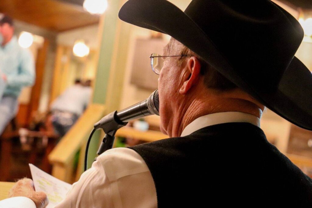A man wearing a black cowboy hat and glasses speaks into a microphone indoors, holding a piece of paper. He is seen from behind, with warm lighting and blurred figures in the background.