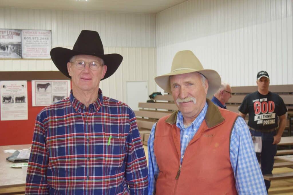 Two older men wearing cowboy hats and casual Western clothing stand indoors in front of wooden bleachers. One wears glasses and a plaid shirt; the other has a mustache and a tan vest. A third man stands in the background.