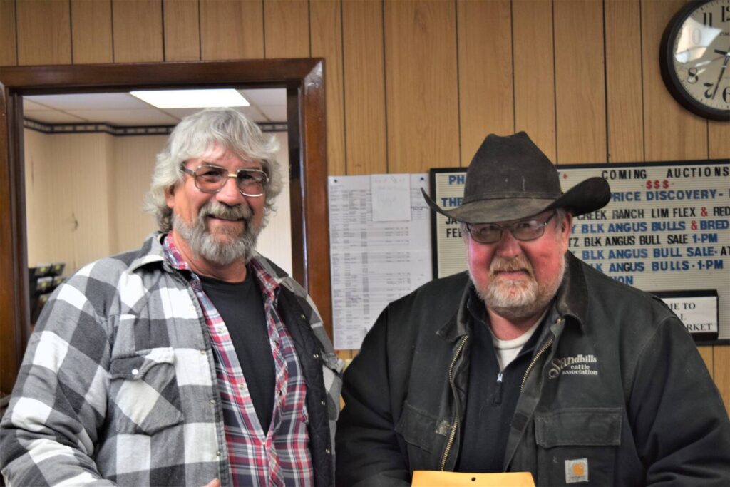 Two older men stand indoors, smiling. One wears glasses, a plaid shirt, and has gray hair and a beard. The other wears a black jacket, glasses, and a black cowboy hat. Wood-paneled walls and a clock are in the background.