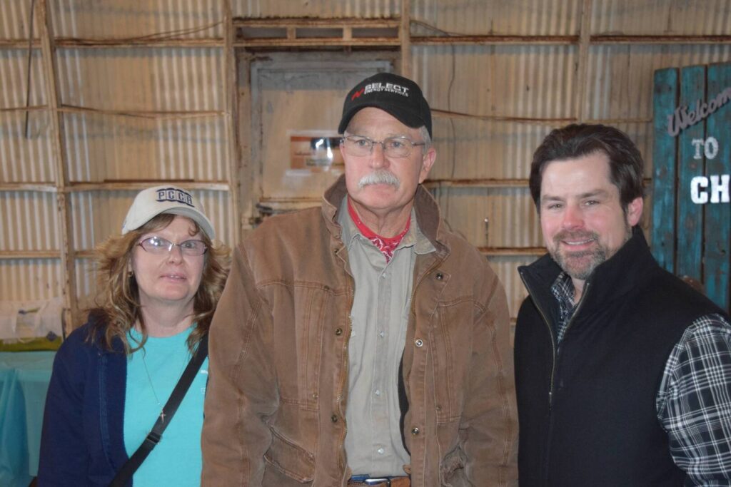 Three adults stand together indoors. The woman on the left wears glasses, a light blue shirt, and a cap. The man in the middle has a mustache, wears glasses, a brown jacket, and a cap. The man on the right wears a black vest and smiles.