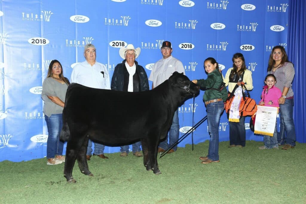 A group of people stand behind a black cow at the 2018 Tulsa State Fair. The backdrop is blue with event logos, and one person holds a ribbon while others smile for the photo.