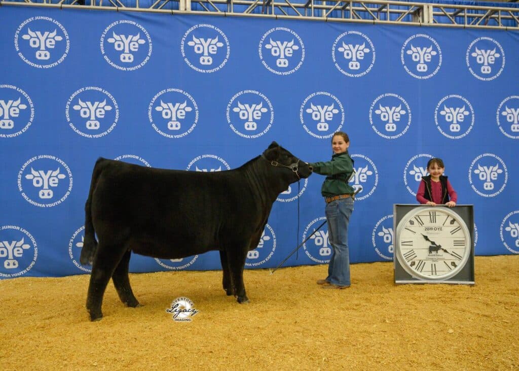 Two girls pose with a black cow at a livestock show. One girl holds the cow’s lead, while the other stands beside a large clock. The background is blue with repeating livestock show logos.