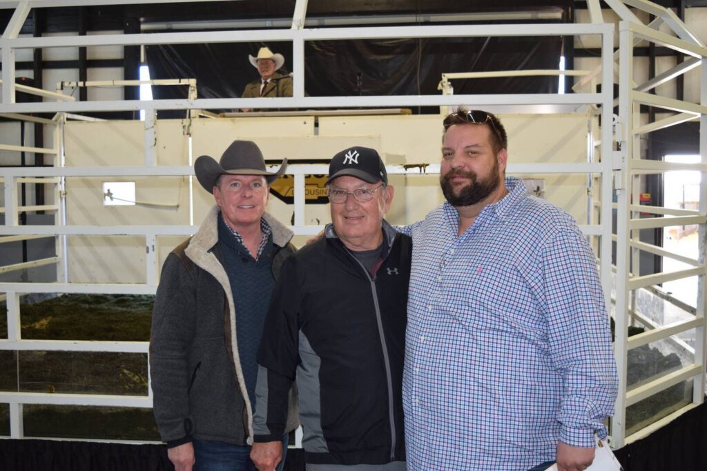 Three men stand together and smile inside a livestock auction ring; one wears a cowboy hat, another a baseball cap, and the third sunglasses on his head. A man in a cowboy hat sits in the background behind the white railings.