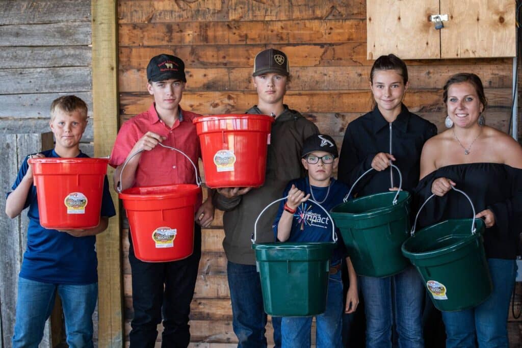 Six people stand in a row against a wooden wall, holding red and green buckets and facing the camera. The group includes boys, one younger child, and a woman, all dressed casually.