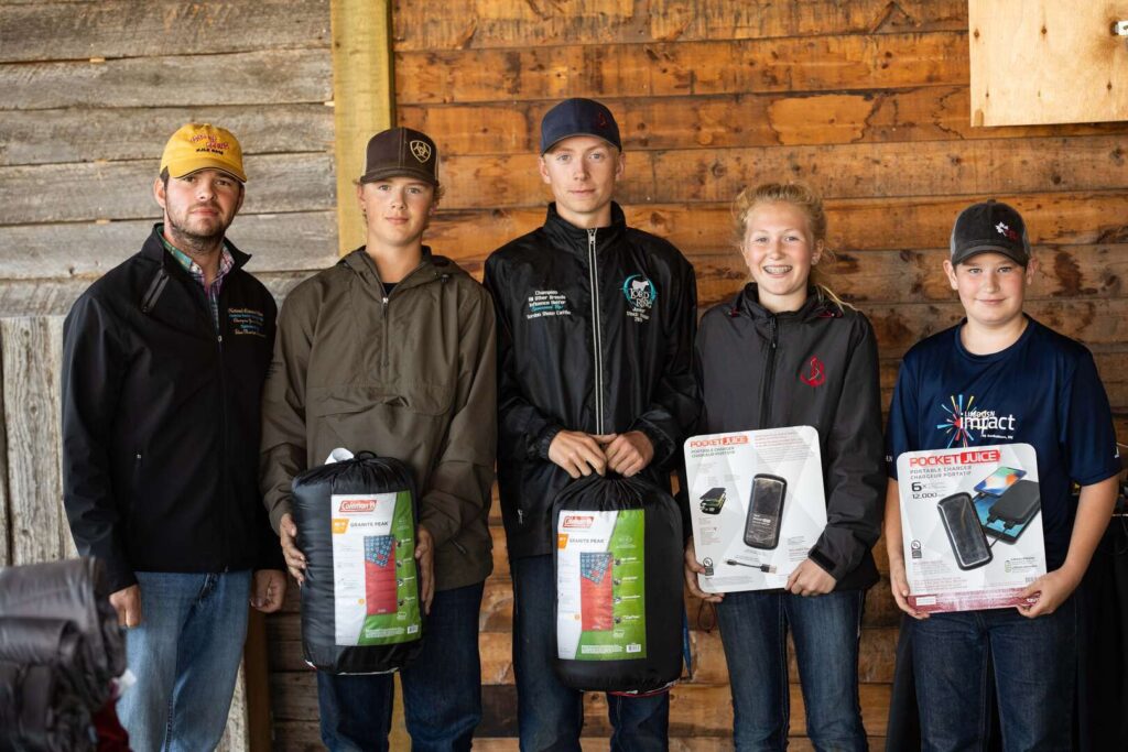 Five young people stand indoors against a wooden wall, holding outdoor gear and awards, such as sleeping bags and lanterns, with a man in a yellow cap standing on the far left. All are smiling at the camera.