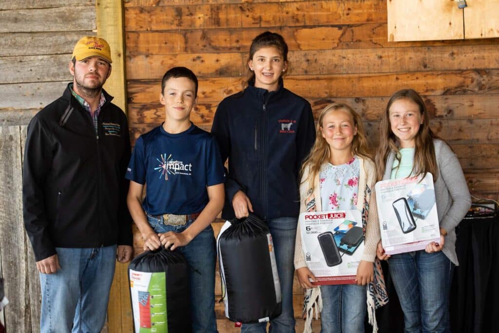 Five people stand together indoors, three holding sleeping bags and two holding boxed portable chargers. They are smiling and casually dressed, with a wooden wall and window in the background.