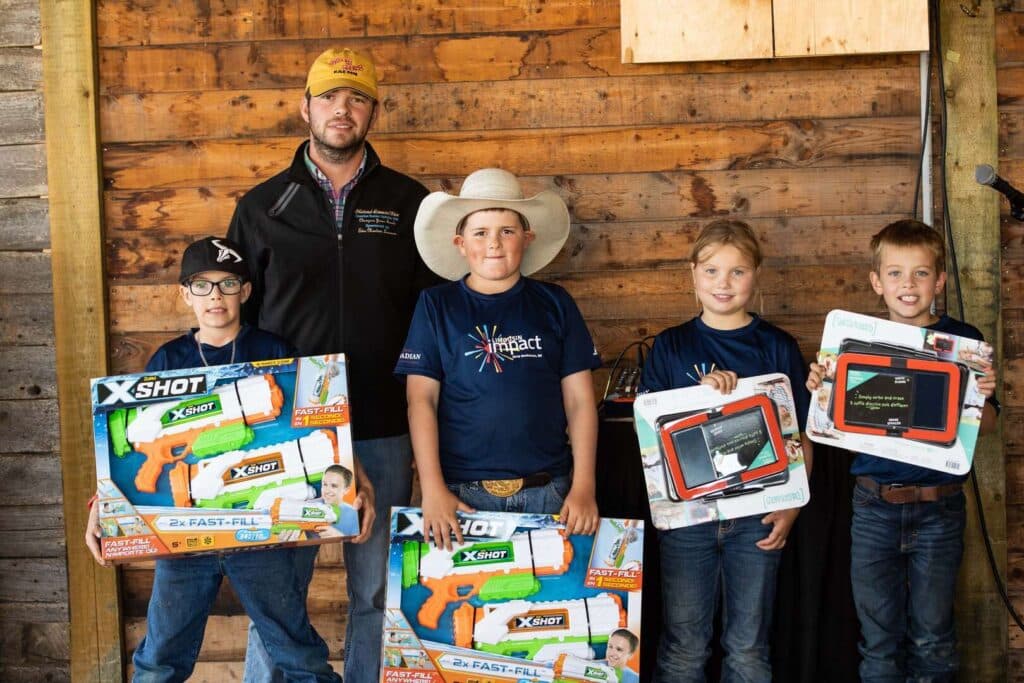 Four children holding toy water guns and tablet boxes stand in front of a wooden wall, smiling. An adult man in a hat and jacket stands behind them.
