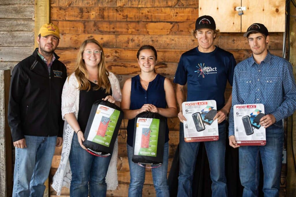 Five people stand indoors in front of a wooden wall, smiling at the camera. Three of them hold packaged sleeping bags, and two hold boxed camping lanterns.