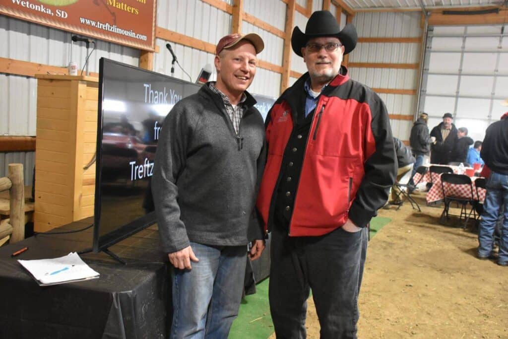 Two men stand smiling indoors at an event, one wearing a gray jacket and red cap, the other in a black cowboy hat and red and black jacket. A large display screen and tables with people are visible in the background.