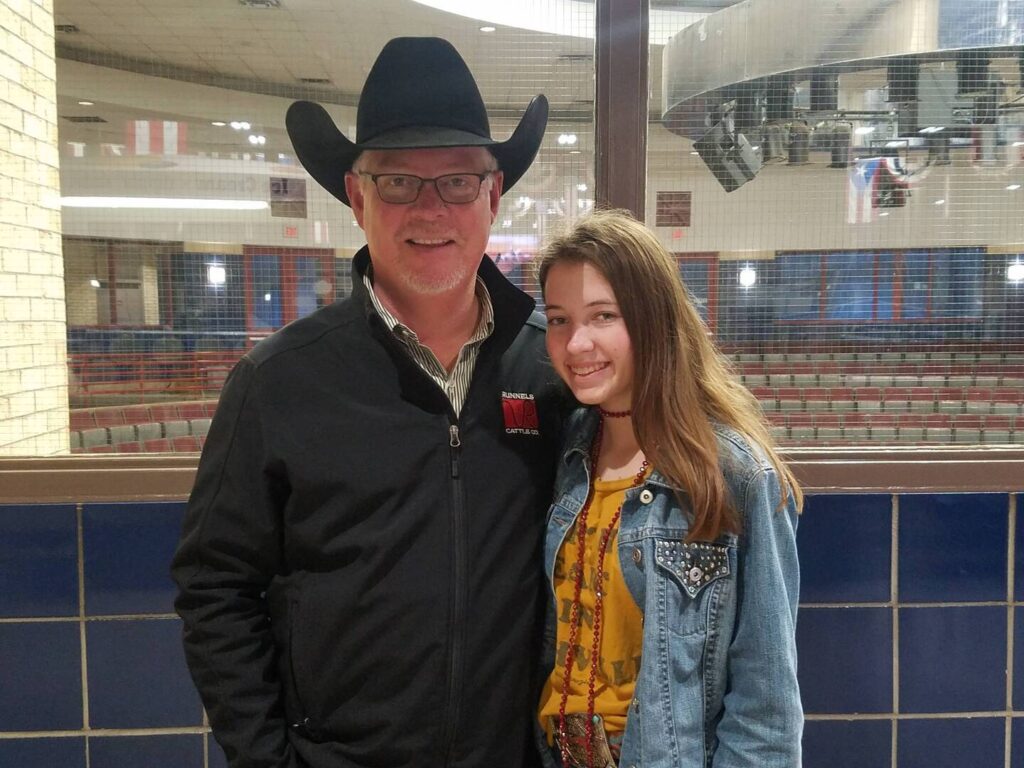 A man in a black cowboy hat and glasses stands next to a smiling teenage girl wearing a denim jacket in a lobby area, with empty auditorium seats visible through the window behind them.