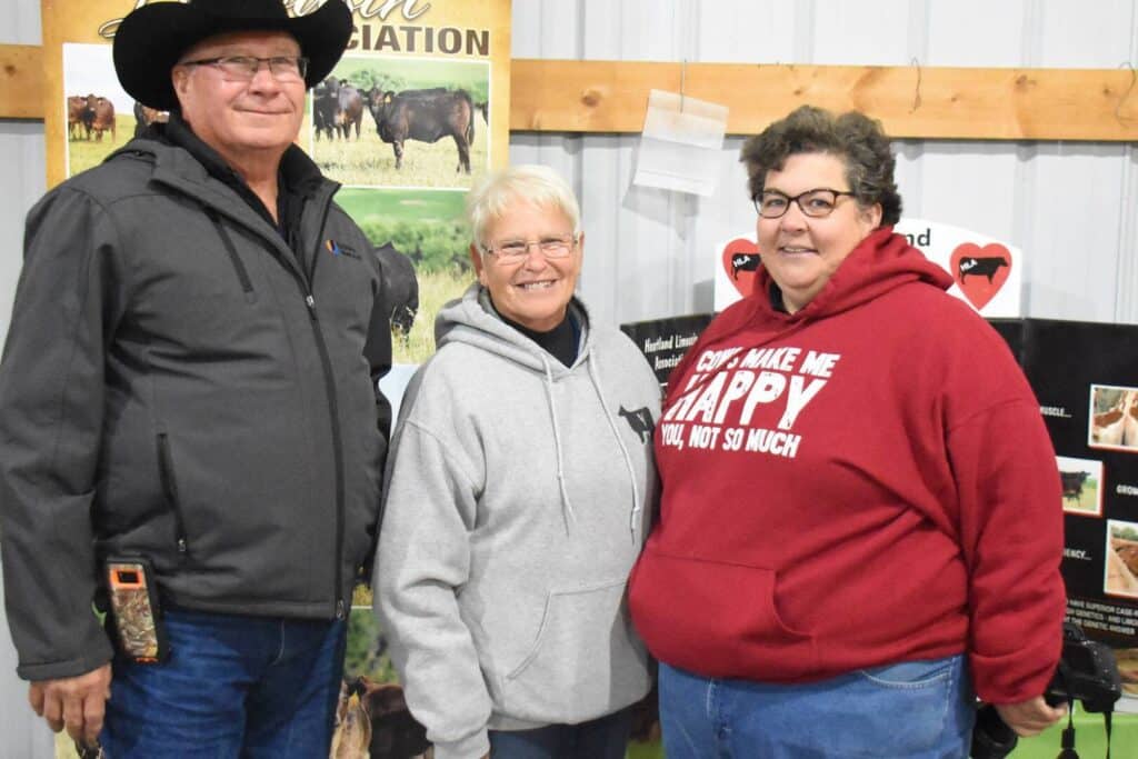 Three people stand indoors, smiling at the camera. One person wears a black cowboy hat and dark jacket, another wears glasses and a gray hoodie, and the third has glasses and a red hoodie with white text. Farm posters are in the background.