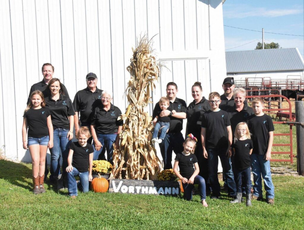 A group of people in matching black shirts pose outdoors beside a white barn, around a tall corn stalk display with a pumpkin and a sign that reads Vorthmann. The group includes adults and children smiling at the camera.