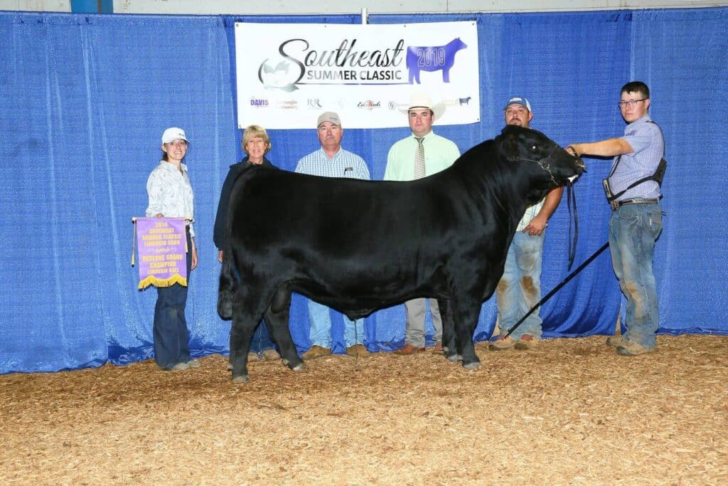 Six people stand around a large black cow at a livestock show, posing for a photo in front of a blue curtain and a “Southeast Summer Classic” banner. One person holds a purple and gold award banner.