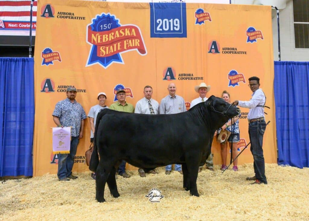 A group of eight people stand behind a black cow at the 2019 Nebraska State Fair, posing for a photo in front of an orange backdrop with the events logo and sponsors.