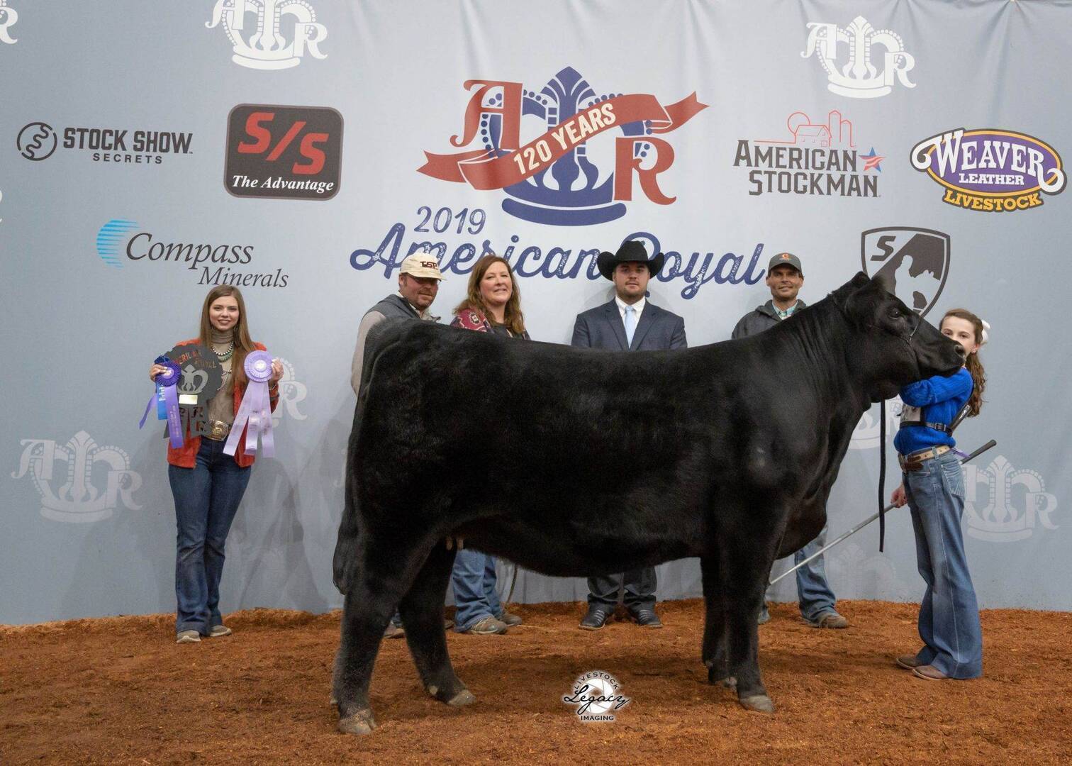Six people stand behind a large black cow at the 2019 American Royal livestock show. Two people hold ribbons, and all are posed in front of a gray backdrop with event logos and sponsors.