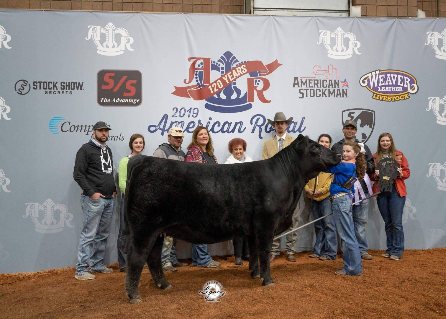 A group of people stand on a red dirt floor behind a large black cow at the 2019 American Royal event. The backdrop displays event sponsors and logos. Some people hold awards and smile at the camera.