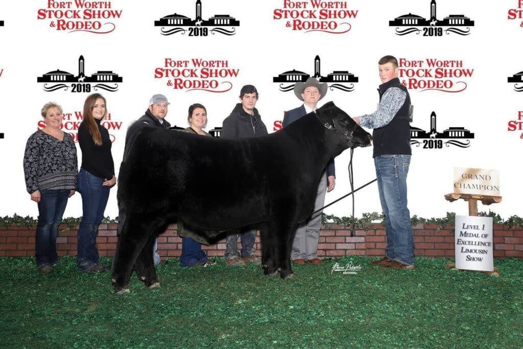 A group of seven people pose with a large black steer in front of a Fort Worth Stock Show & Rodeo 2019 backdrop. A sign nearby reads Grand Champion Level 1 Medal of Excellence Limousin Show.