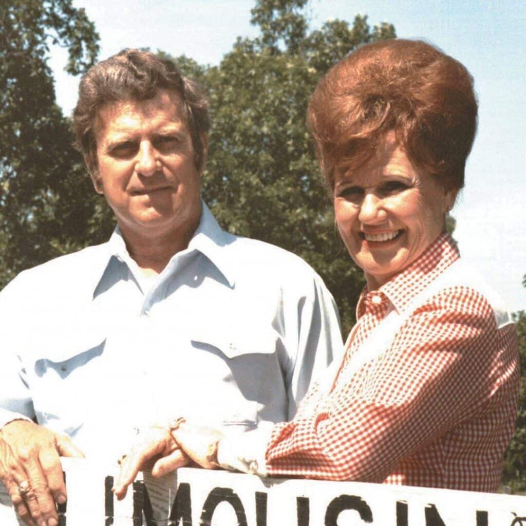 A man in a light blue shirt and a woman in a red checked blouse stand outdoors, smiling, with trees in the background and part of a sign visible at the bottom of the image.