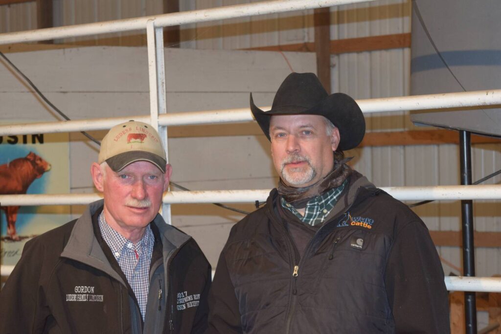 Two men wearing jackets and hats stand indoors in front of a white metal fence. One man wears a beige cap and the other a black cowboy hat. Both are looking at the camera; there is a poster and barn walls in the background.