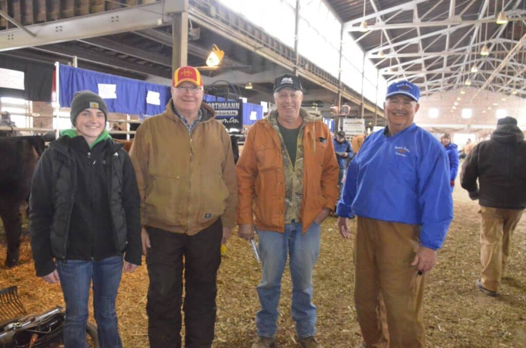 Four people stand indoors at a livestock event, smiling at the camera. They are dressed in casual outdoor clothing, including jackets and hats. Cattle pens and event banners are visible in the background.