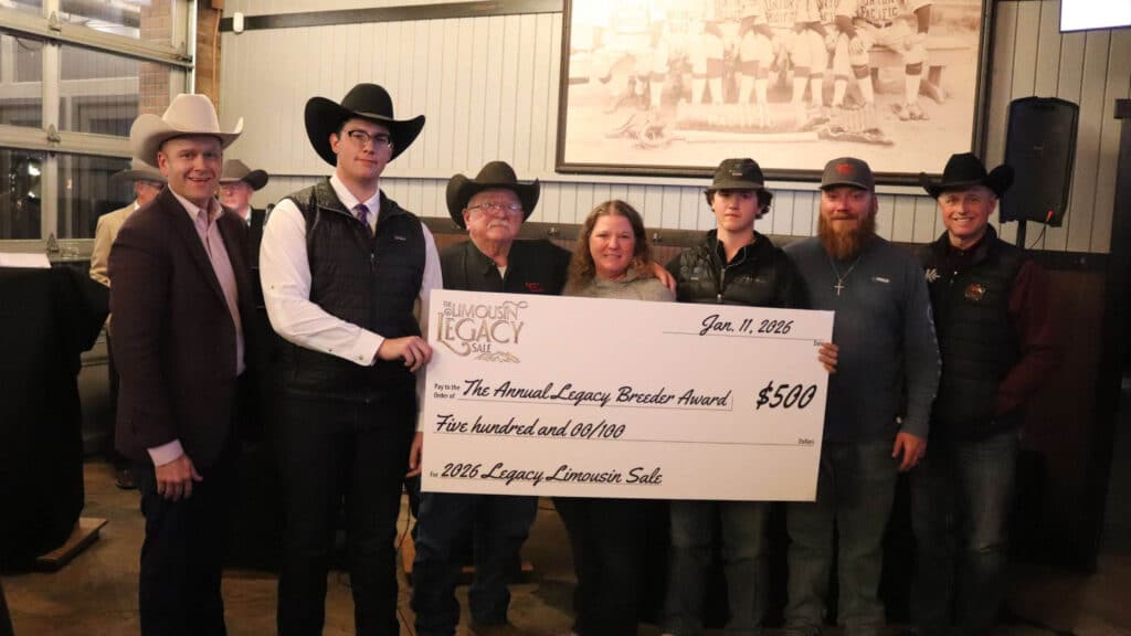 Seven people wearing cowboy hats and western attire stand indoors, smiling and posing with an oversized $500 check for the 2025 Legacy Breeder Award at the 2025 Legacy Limousin Sale.