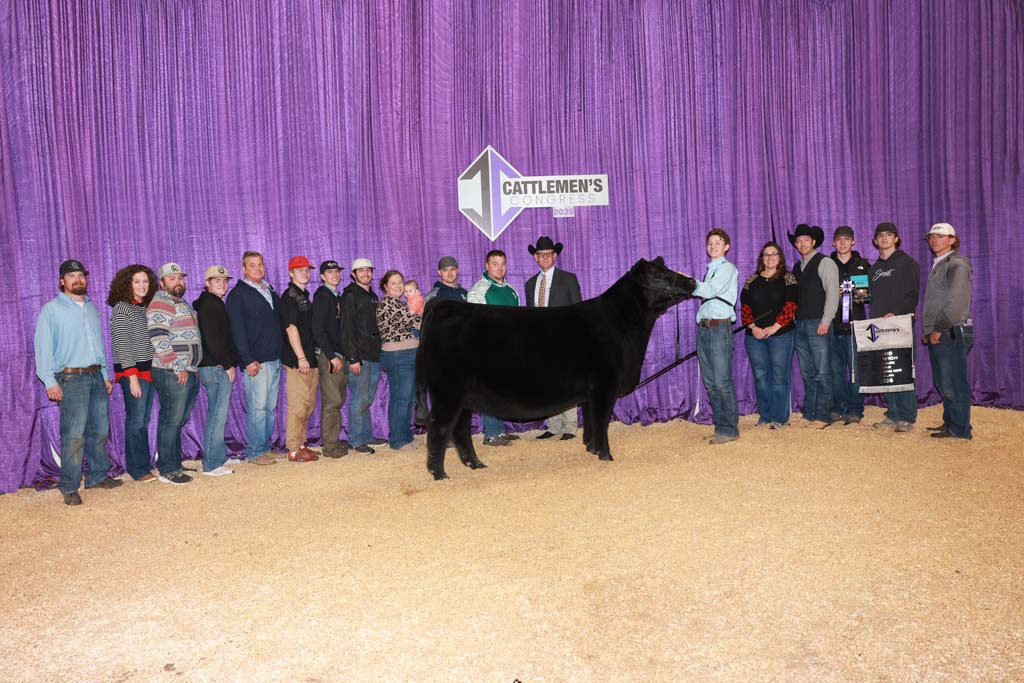 A group of people stands in a row behind a black cow at the Cattlemen's Congress, with a purple curtain and event sign in the background.