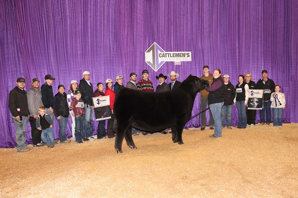 A group of people stand behind a black cow at a livestock show in front of a purple curtain displaying a sign that reads "Cattlemen's Congress.
