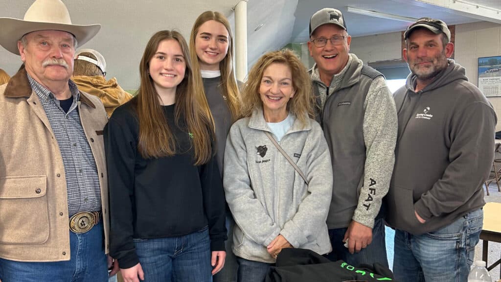 A group of six people—three men, two young women, and one older woman—stand close together indoors, smiling at the camera. One man wears a cowboy hat and others wear casual or outdoor clothing.