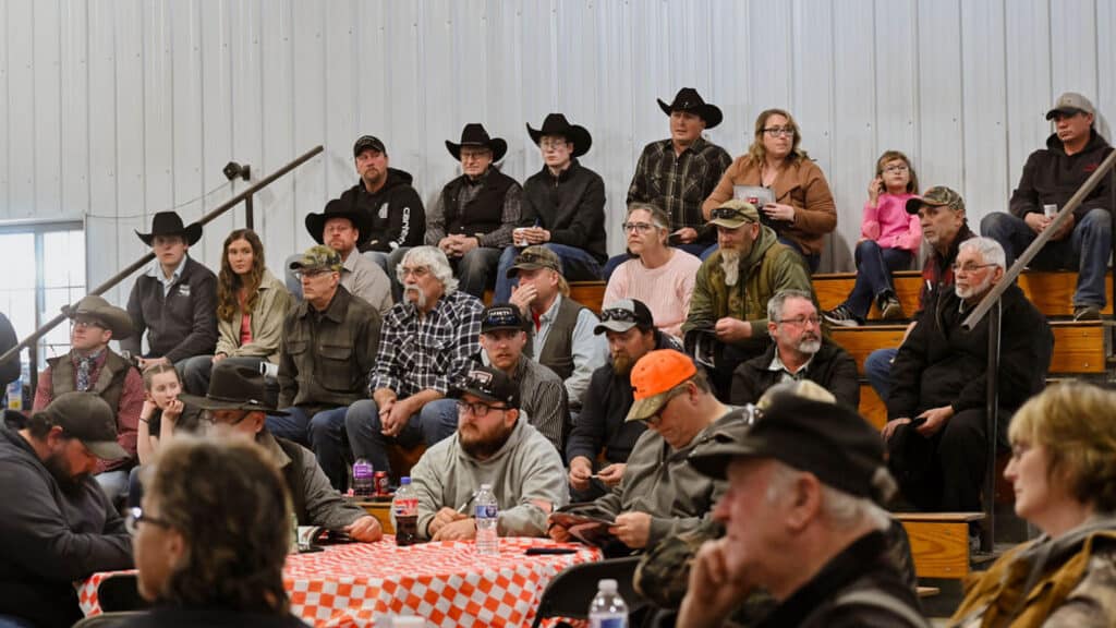A group of people, many wearing cowboy hats and casual clothing, sit on bleachers in a metal-walled indoor space, watching an event. Some sit at nearby tables with checkered tablecloths.