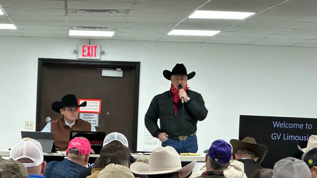 A man in a black cowboy hat and red bandana speaks into a microphone at an indoor event, while another man in a cowboy hat sits at a desk beside him. Audience members in cowboy hats face the speakers.