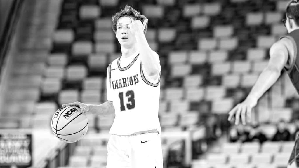 A basketball player in a Warriors jersey with the number 13 dribbles an NCAA basketball on the court, while an opposing player reaches out towards him. The seats in the background are mostly empty.