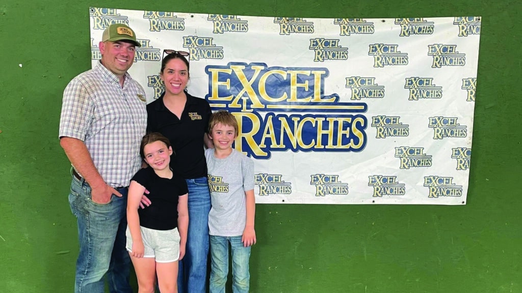 A smiling family of four stands in front of a banner that reads Excel Ranches multiple times. Two adults and two children pose together against a green wall.