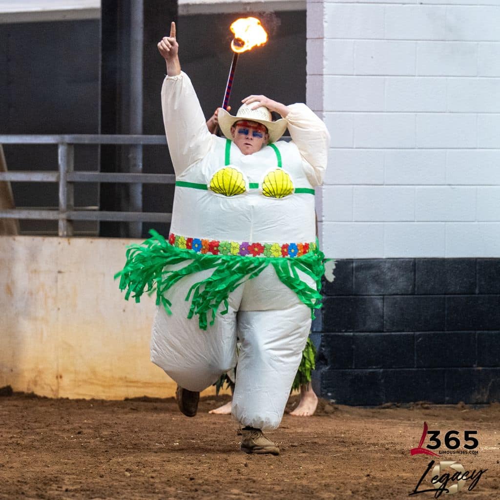 A person in an inflatable hula dancer costume with seashells and a green grass skirt runs indoors, holding a flaming torch and pointing upward, in what appears to be a rodeo or arena setting.