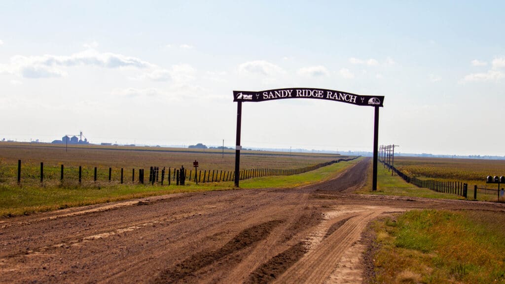 A dirt road leads to a wide rural landscape, passing under a metal archway sign that reads Sandy Ridge Ranch. Fences and open fields stretch into the distance under a partly cloudy sky.
