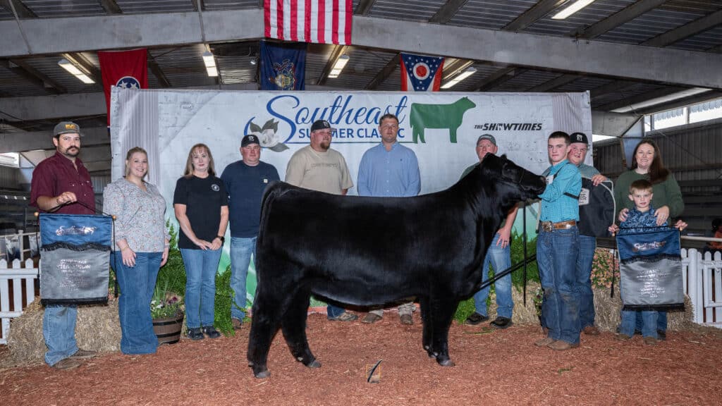 A group of people pose indoors with a black cow at a livestock event. Two people hold award banners. A large Southeast Summer Classic banner is displayed in the background. Flags hang above them.