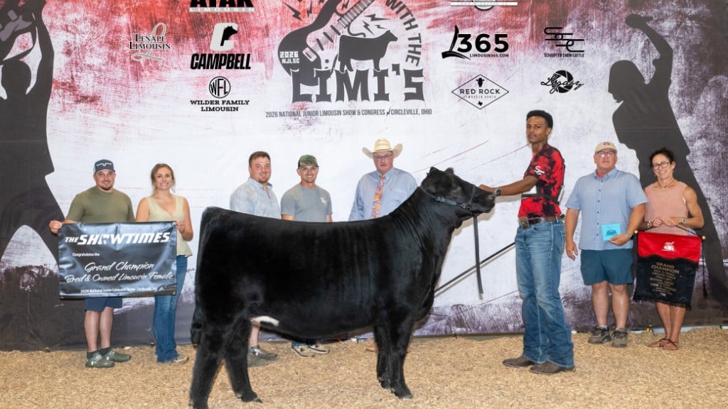 A group stands at a livestock show with a black cow in the center. One man holds the cow by a lead rope. Others display banners, awards, and ribbons. The backdrop features sponsor logos and event branding.