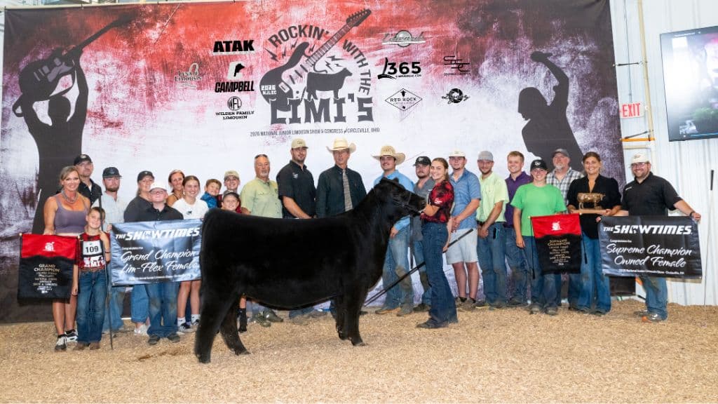 A group of people pose with a black cow and award banners in front of a red and white backdrop at a livestock show event.