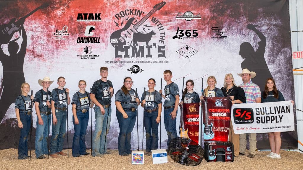 A group of young people in matching shirts pose with adults in front of a “Rockin’ With the Cattle” backdrop, holding awards and a S/S Sullivan Supply banner at a livestock event.