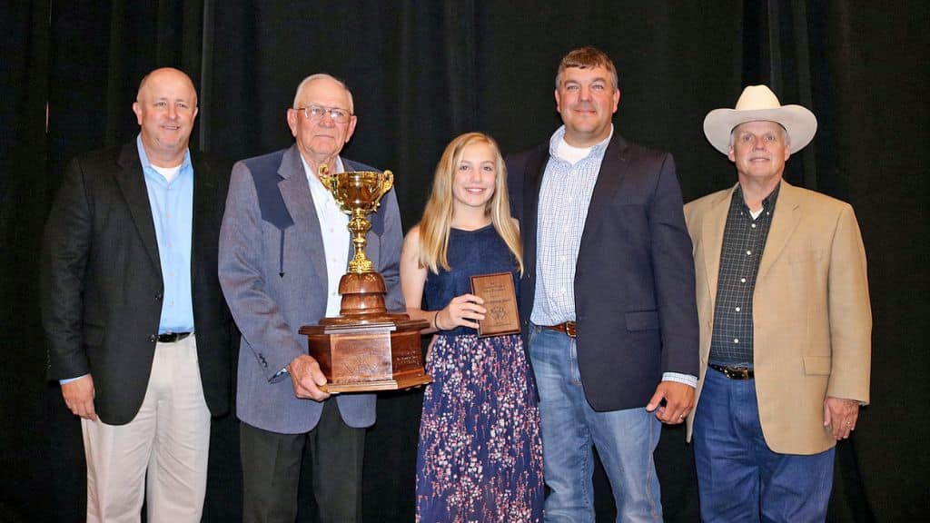 Five people stand together, dressed in semi-formal attire. The man second from the left holds a large trophy while a young woman in the center holds a plaque. They are posing in front of a black backdrop.