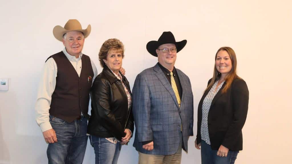 Four adults stand side by side against a plain wall; two men wear cowboy hats, one woman wears a leather jacket, and another woman wears a blazer. All are smiling and dressed in business-casual attire.