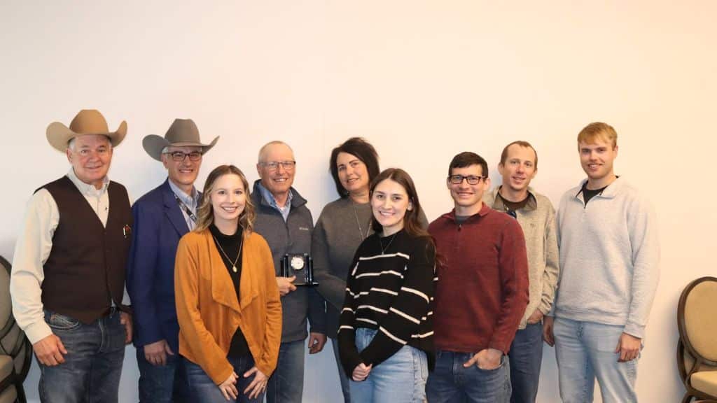 Nine people pose indoors against a plain wall, with some smiling and one holding a small award. Two men on the left wear cowboy hats and western attire, while the others are dressed in casual and business-casual clothing.