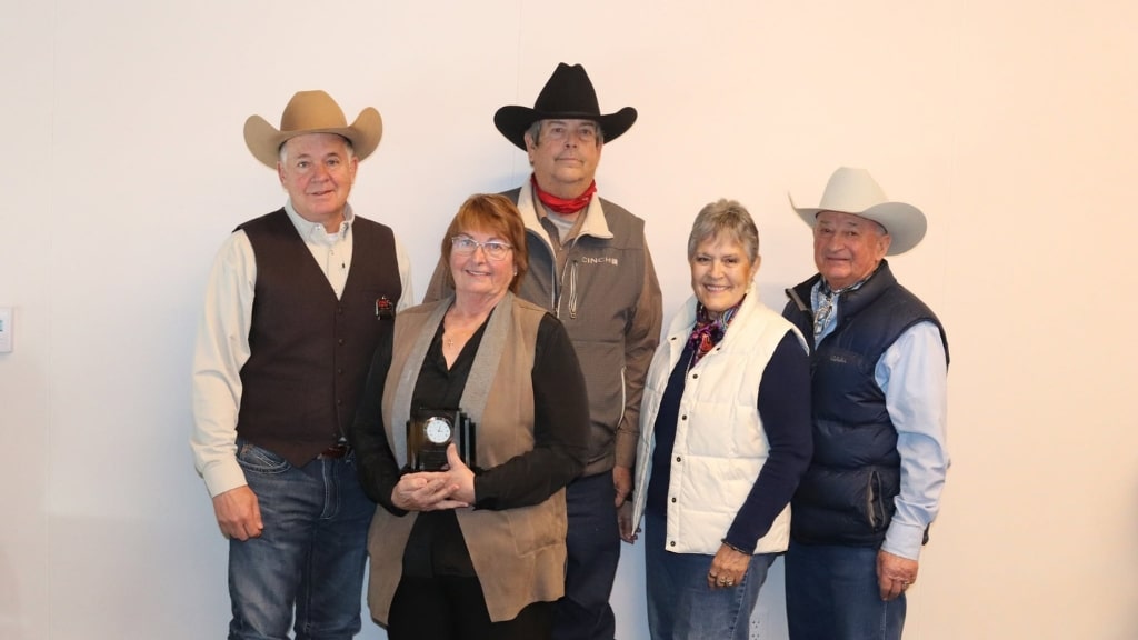 Five older adults wearing cowboy hats and western-style clothing pose in front of a plain light-colored wall. One woman in front holds a round award. All are smiling and standing closely together.